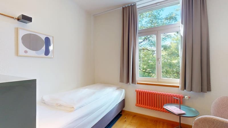 a small bed, single bed, bed linen, window with curtain, small chair, night lamp, radiator, small side table, picture frame on wall