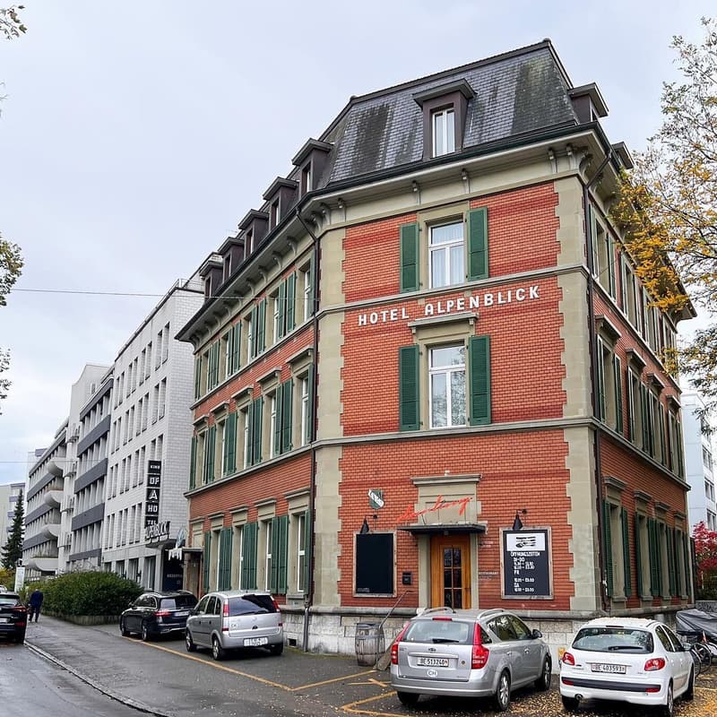 The building has a red brick facade with green shutters, a gray roof, and a sign that reads 'Hotel Alpenblick' above the entrance. Several cars are parked in front of the building, and a person is walking along the sidewalk.