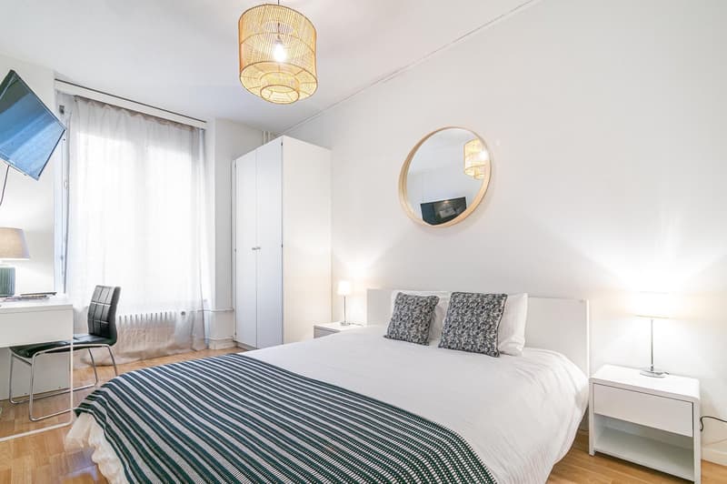 white walls, hardwood floor, bed, side tables, lamps, desk, chair, round mirror, television, wardrobe
