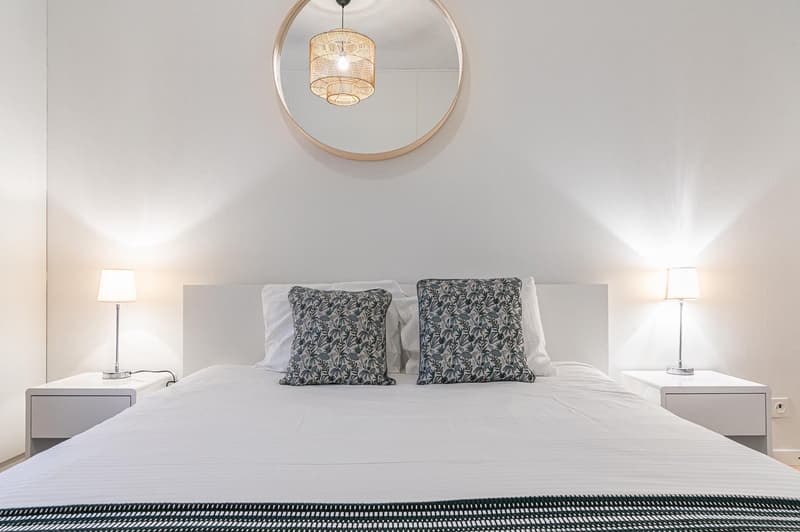 A white bed with white pillows and gray sheets, lamps on the nightstands, a white ceiling, a round mirror on the wall