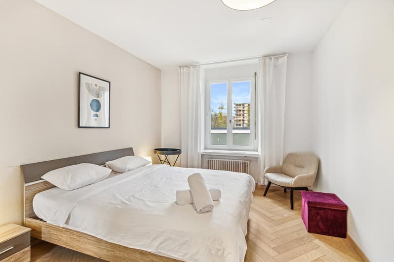 a bed with white linens, two nightstands, a picture on the wall, a chair near the window, a round ottoman, a small table