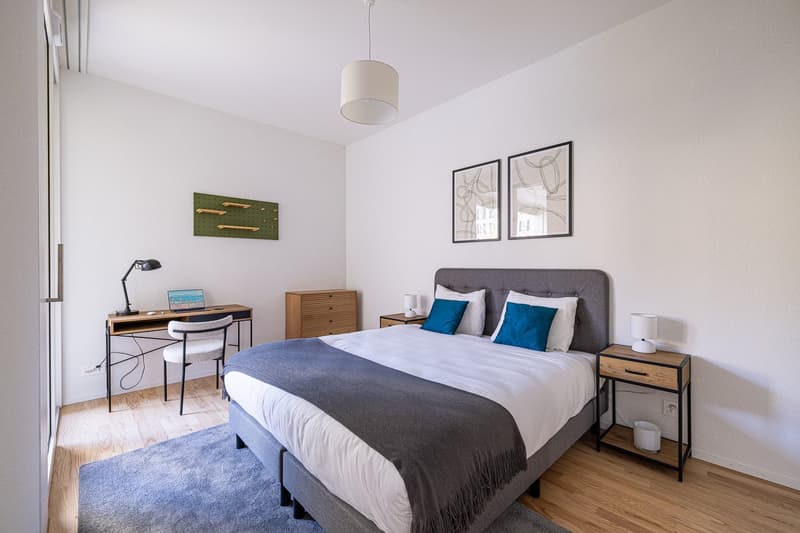 Bedroom with bed, desk, chair, nightstands, lamps, rug, and framed pictures on the wall.