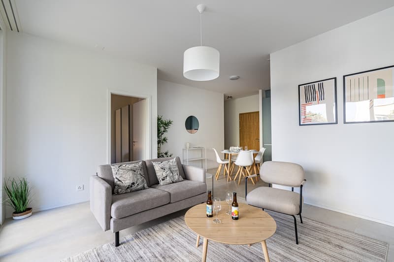 modern living room with gray sofa, wooden coffee table, round mirror, white walls, and a dining area with a table and chairs