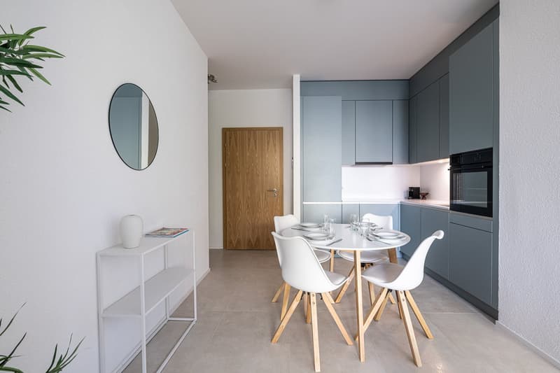 Dining area with a round table and chairs, white shelves, wooden door, round mirror, and a kitchen with an oven and cabinets