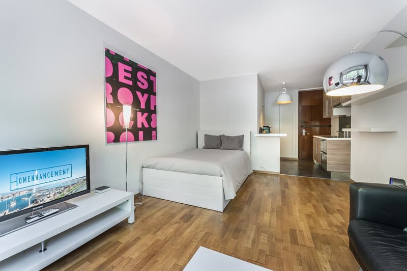 small bedroom with bed, TV, lamp, and a small kitchen area