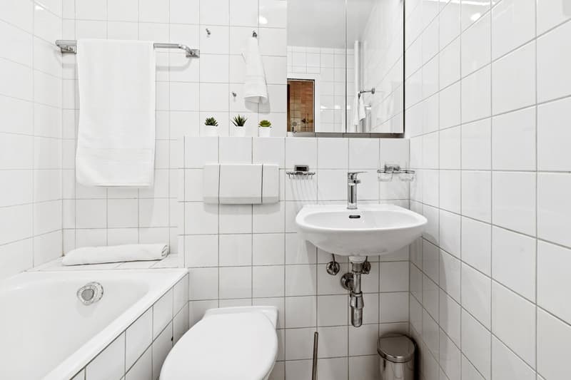 White tiled bathroom with a toilet, sink, and towel rack. There is a small shelf with potted plants above the sink. The bathroom appears to be clean and minimalist in design.
