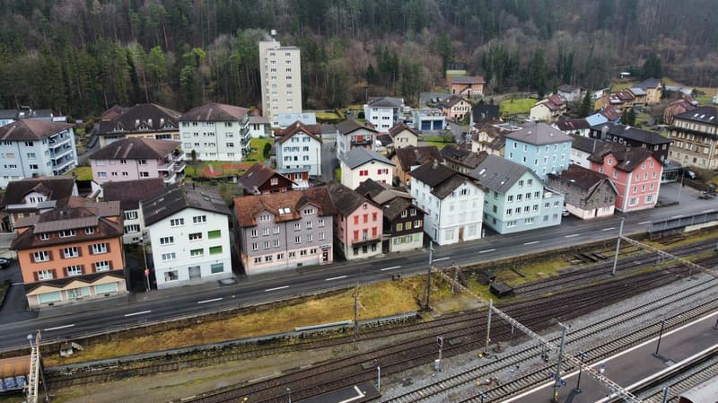 Various residential buildings, mostly multi-story, near railway tracks with an aerial view, surrounded by trees and small hills