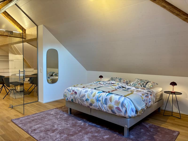 A bedroom with wooden floor, a large bed, a mirror, bedside tables, lamps, and a glass wall with a view of a dining table and chairs.