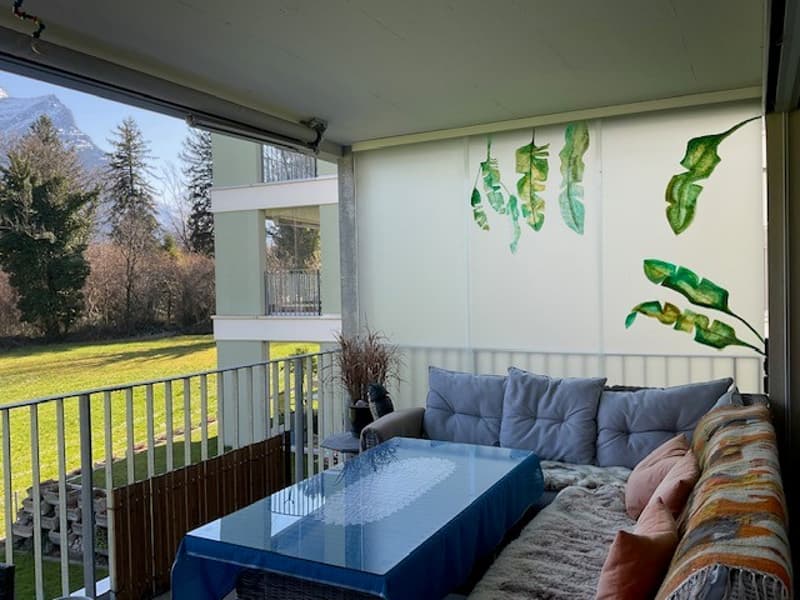 Furnished balcony with couch, glass table, green plants, decorative wall art, mountain view