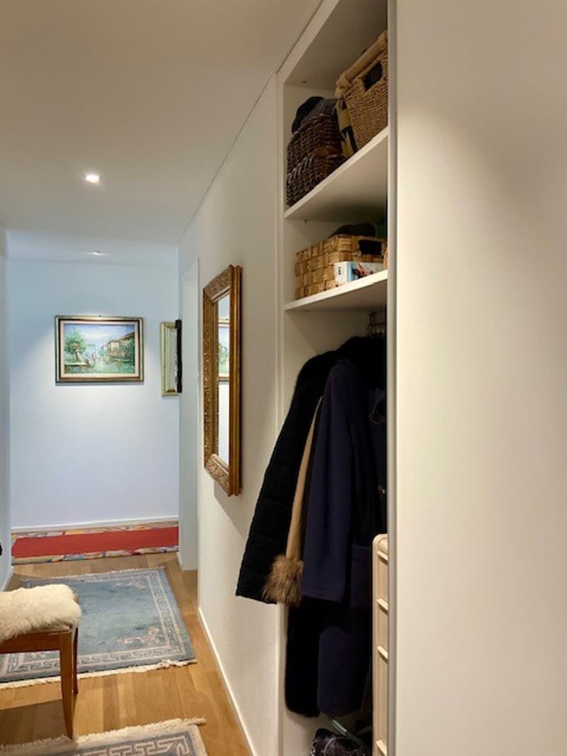 A narrow hallway with wooden floor, white walls, and built-in wardrobe. There are two small rugs, a chair with a fur seat, and a picture frame on the wall. There are shelves with baskets and coats, and a mirror is mounted on the wall.