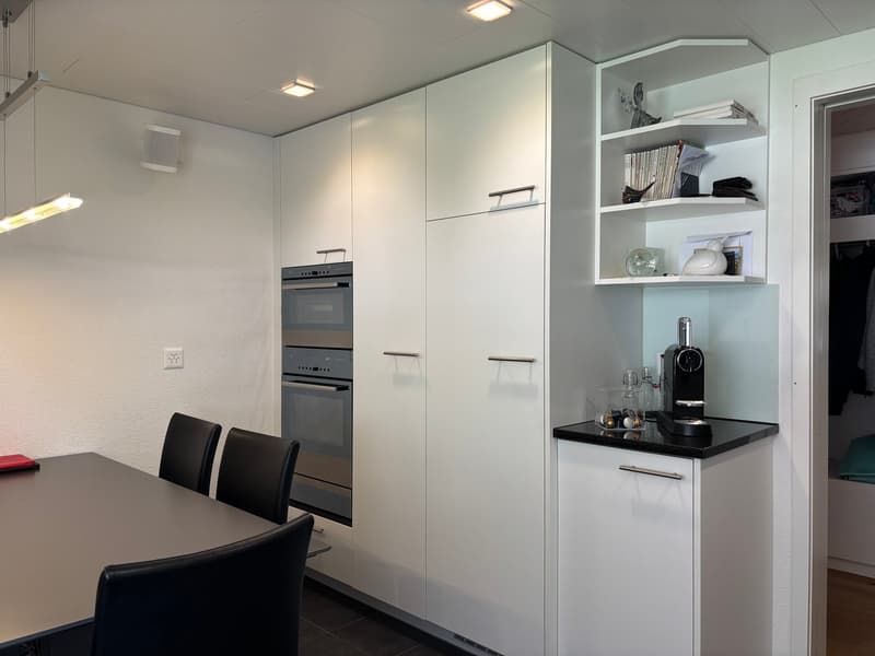 modern kitchen with white cabinets, black countertops, built-in oven, dishwasher, coffee machine, dining table, and chairs