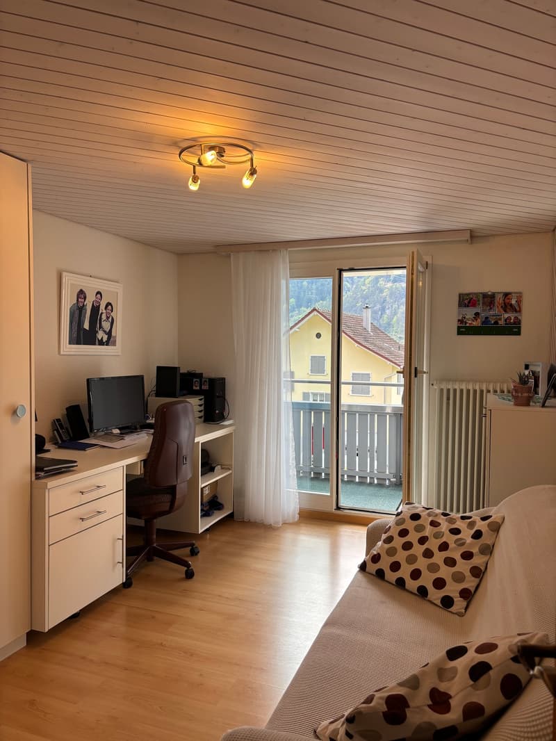 Desk with computer, chair, cabinet, couch with pillows, radiator, picture frame, balcony with railing, window with curtain