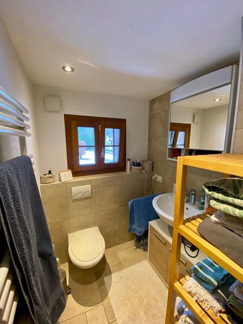 tiled walls, window with wooden frame, white toilet, white sink, mirror, wooden shelf with towels, radiator