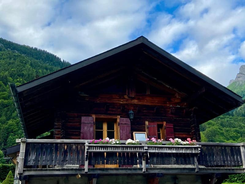 Alpine-style house with wooden facade, steeply pitched roof, three windows, wooden balcony with flower boxes, set against a mountainous backdrop