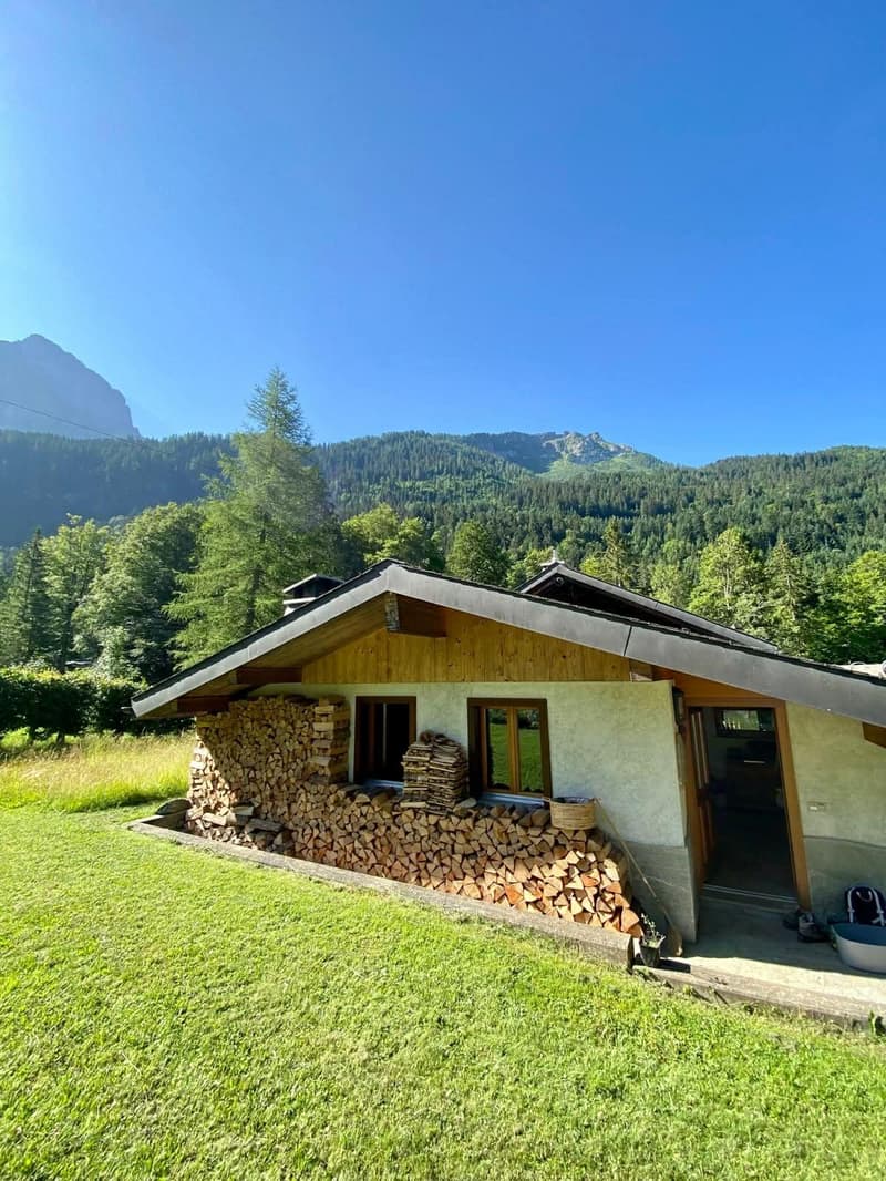 Wood and white house with stack of wood in front, surrounded by trees, mountains in the background.
