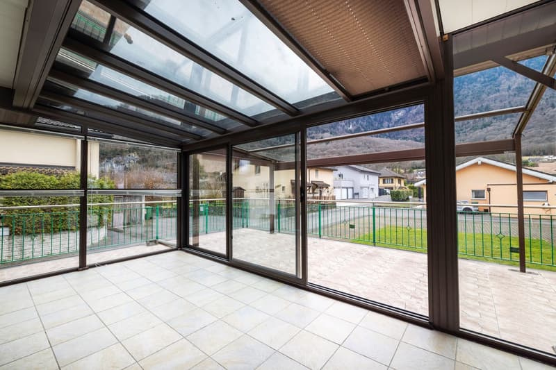 Large glass windows and sliding doors, wooden ceiling, tiled floor, balcony with railing overlooking a residential area and mountains in the background