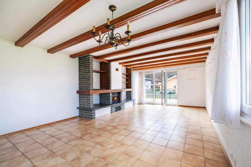 Spacious living room with exposed wooden beams on the ceiling, a brick fireplace, and sliding glass doors leading to an outdoor area. The room has tile flooring and a chandelier lighting fixture.