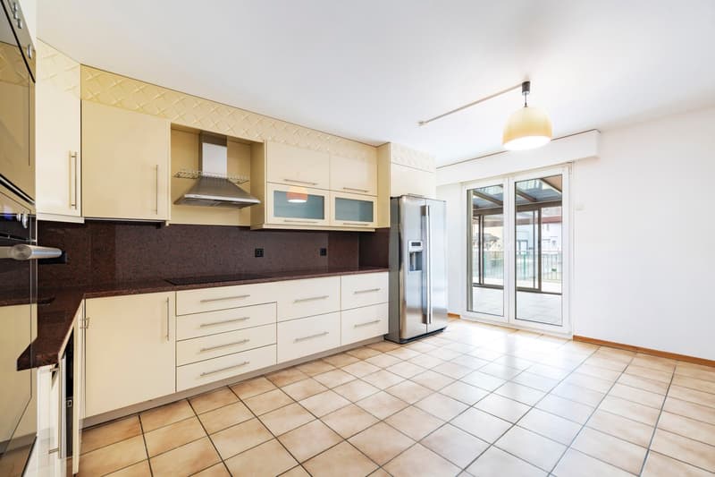 Spacious kitchen with beige cabinets, black countertops, tiled flooring, and a stainless steel refrigerator. The kitchen has a range hood and appears to have a dishwasher installed.