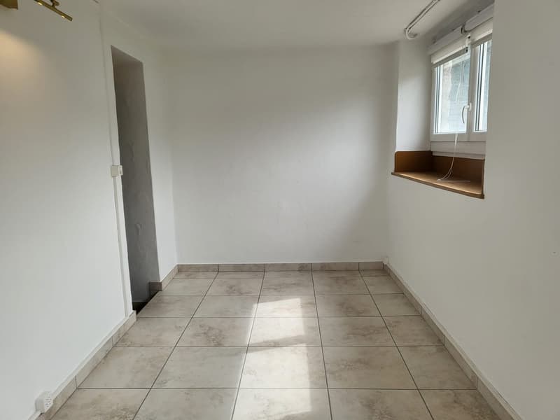 This image shows a small, empty room with white walls and a tiled floor. There is a window on one wall and a built-in shelf or ledge along the bottom of the window. The room appears to be a simple, minimalist space.