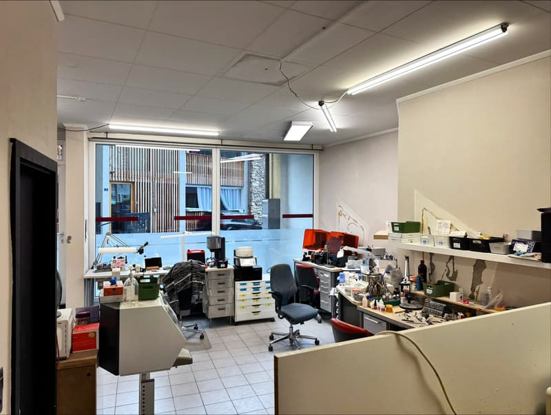Office space with desks, chairs, cabinets, shelves, and equipment. Large glass windows with blinds. White ceiling with fluorescent lights.
