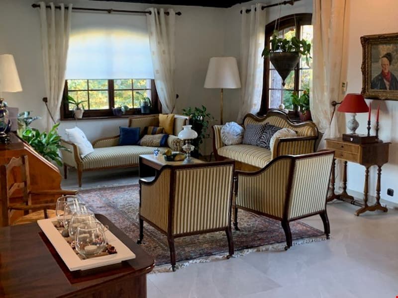 Spacious living room with large windows, wooden furniture including a sofa, armchairs, and coffee table, decorative items like lamps and plants, and a fireplace.