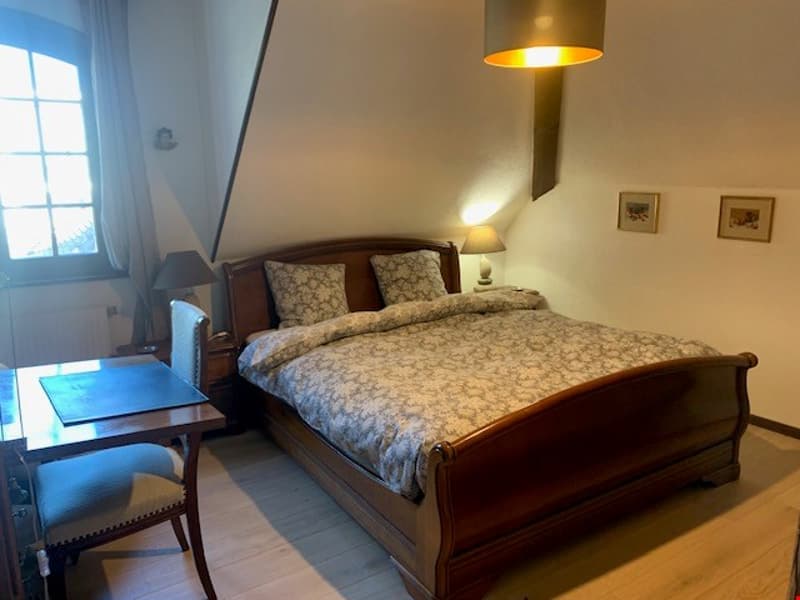Spacious bedroom with a wooden sleigh bed, patterned bedding, a wooden side table, and a floor lamp. The room has a large window that provides natural light.