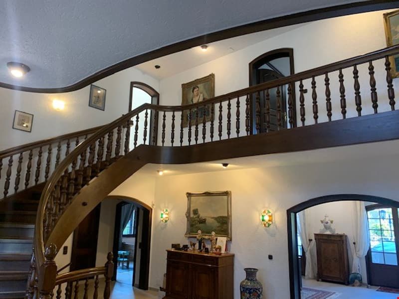 Spacious two-story foyer with a curved wooden staircase, ornate wooden railings, and arched doorways. The walls are painted in a light color, and there are decorative mirrors and artwork hung on the walls. The space appears to be part of a larger resident