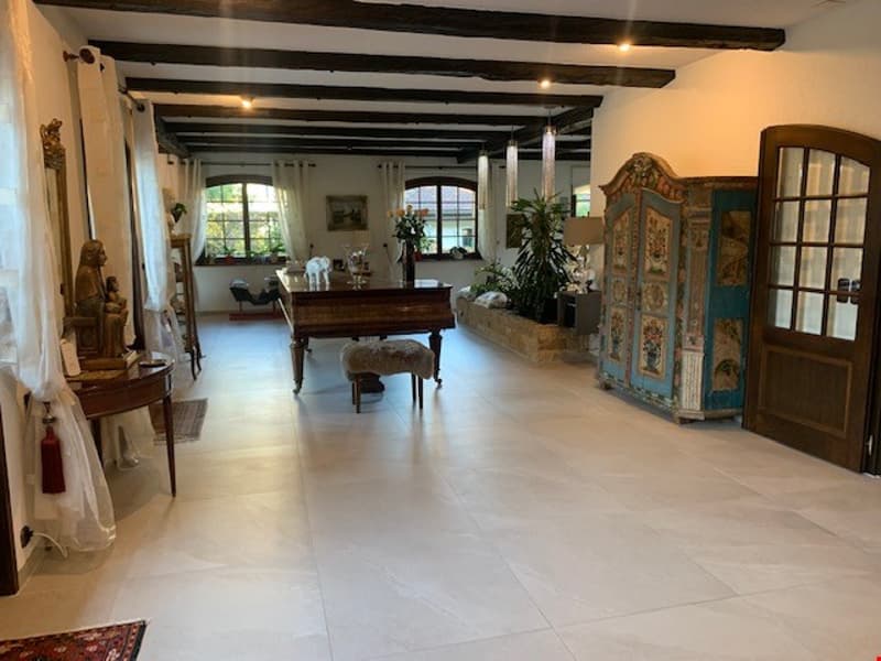 Large open-concept living space with hardwood floors, exposed wooden beams, and a fireplace. The room features a grand piano, antique furniture, and decorative elements like a screen and potted plants.