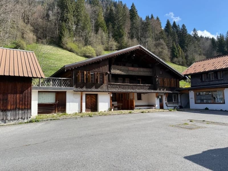 A traditional wooden chalet-style building with a sloped roof, located in a rural, mountainous area surrounded by pine trees. The building has a garage and appears to be part of a larger complex or village.