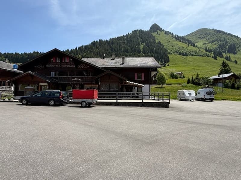 A traditional Swiss mountain village with wooden chalets, surrounded by lush green hills and mountains. There is a parking area in front of the buildings, and some vehicles and trailers are visible.