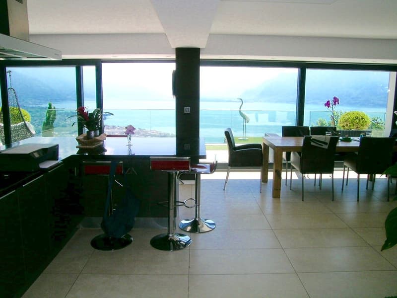 Spacious open-plan living area with large windows offering a scenic view of the lake or ocean. Modern kitchen with bar stools, dining table and chairs, and decorative plants and flowers. Sleek and minimalist interior design with tile flooring.