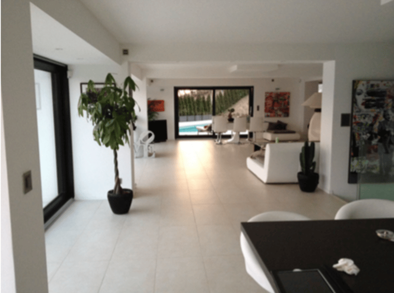 Open-concept living space with white walls, tile flooring, large windows, and a potted plant. The space appears to lead out to a balcony or terrace area.
