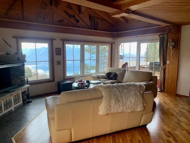 Spacious living room with large windows offering a scenic view, wooden beams on the ceiling, hardwood flooring, comfortable leather sofas, and a fireplace.