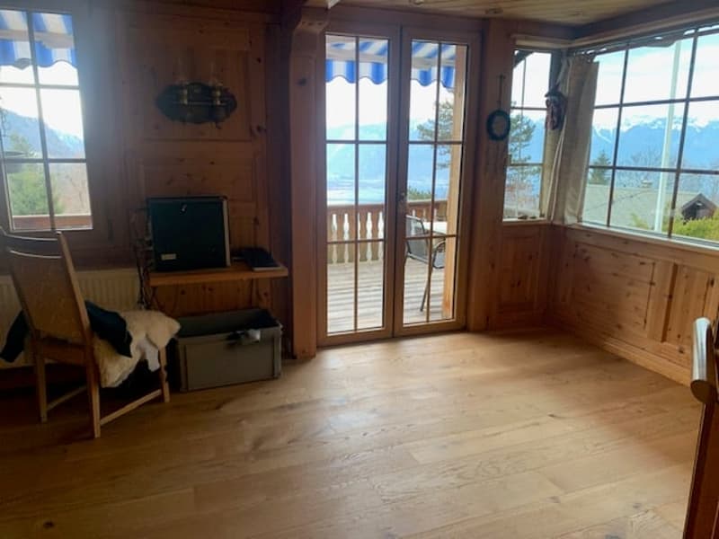 Wooden interior, large windows with a view, wooden floor, TV stand, and a balcony or terrace visible through the glass doors.