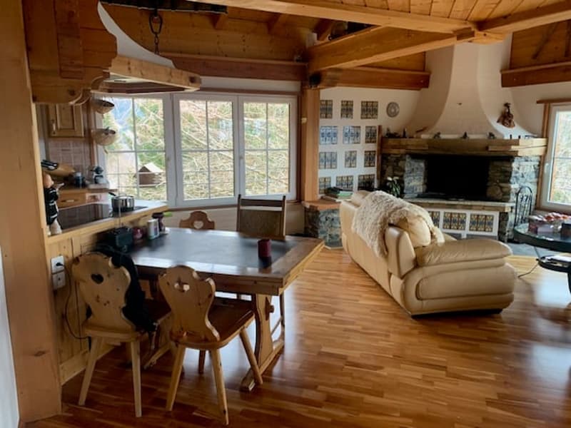Rustic cabin-style interior with wooden beams, large windows, a fireplace, a dining table with wooden chairs, and a cozy sofa