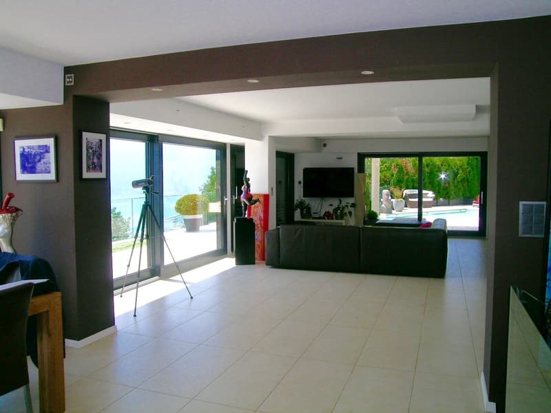 large living room, white tiles, large windows, sliding glass doors, black leather couch, TV, tripod, plants, pool view