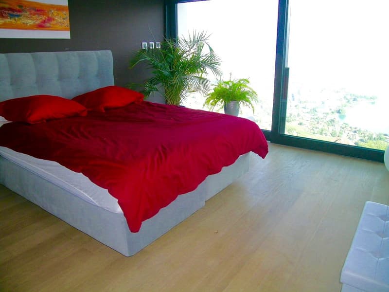 Bedroom with a double bed, red bedspread, two red pillows, wooden floor, large windows, plants, city view