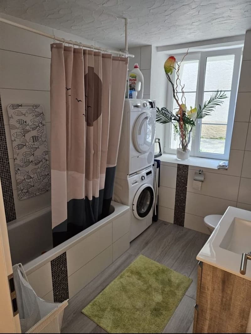 Bathroom with bathtub, sink, toilet, white tiled walls, wooden floor, shower curtain, washing machine on top of another washing machine, small rug, potted plant on window sill, and towel.