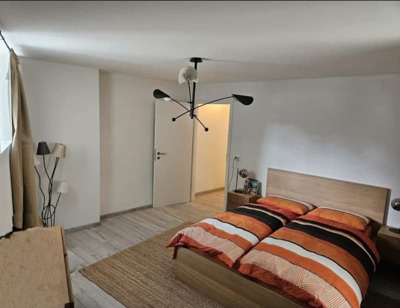 spacious bedroom with white walls, wooden floor, a large bed with orange and black striped bedding, a wooden nightstand, and a unique chandelier