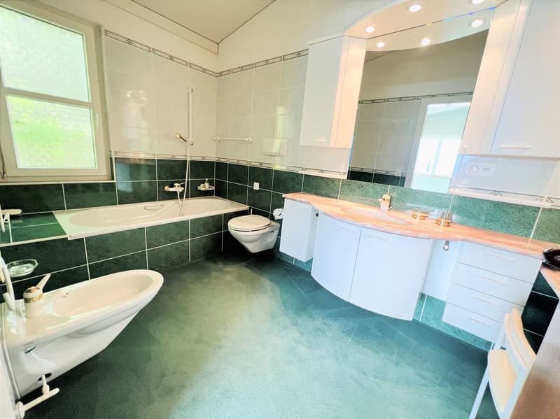 Large bathroom with green and white tile, freestanding bathtub, vanity with countertop, toilet, and large window