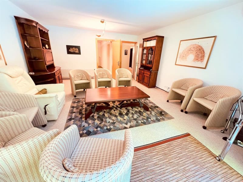 The image shows a spacious living room with a wooden dining table and chairs, a wooden cabinet, and a patterned area rug. The room has a cozy and traditional feel, with plaid and striped upholstered furniture. The walls are painted a light color, and ther