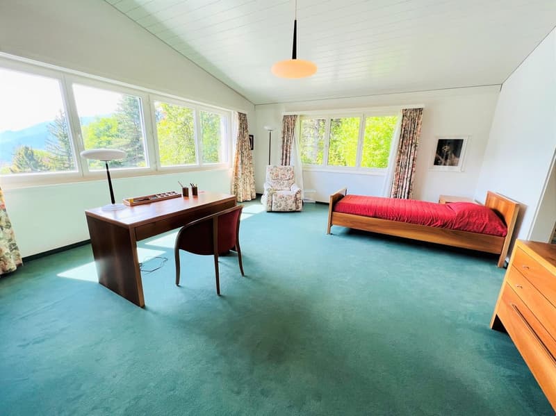 Spacious bedroom with large windows providing a scenic view, wooden desk and chair, red bed, dresser, and floral curtains
