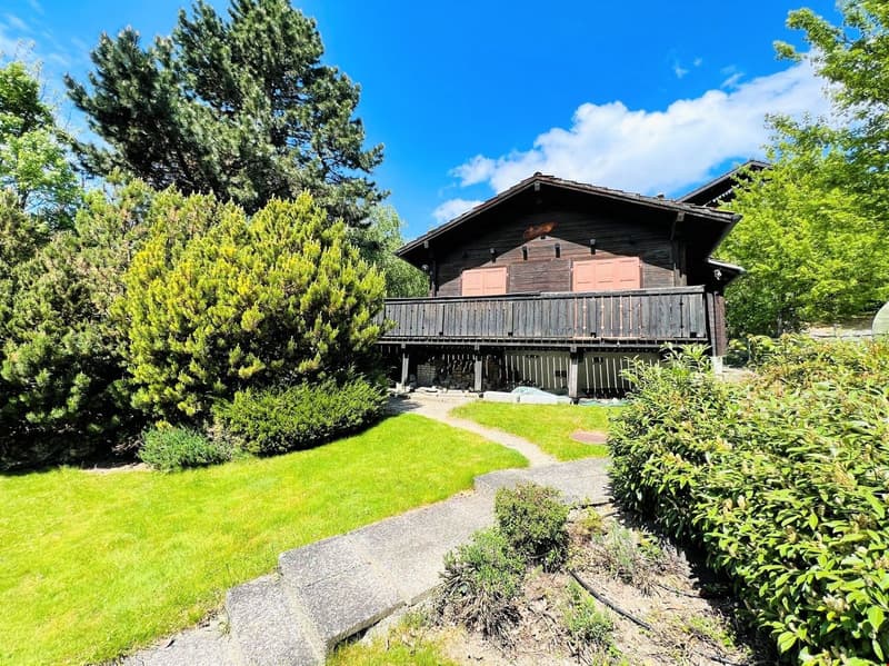 Two-story wooden cabin with a balcony, surrounded by lush greenery and pine trees. The cabin has a rustic, traditional design with a large balcony overlooking a well-maintained lawn and garden area.