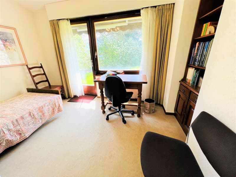 Bedroom with a desk, chair, and bookshelves. Large window overlooking a balcony or terrace with a view of greenery outside.