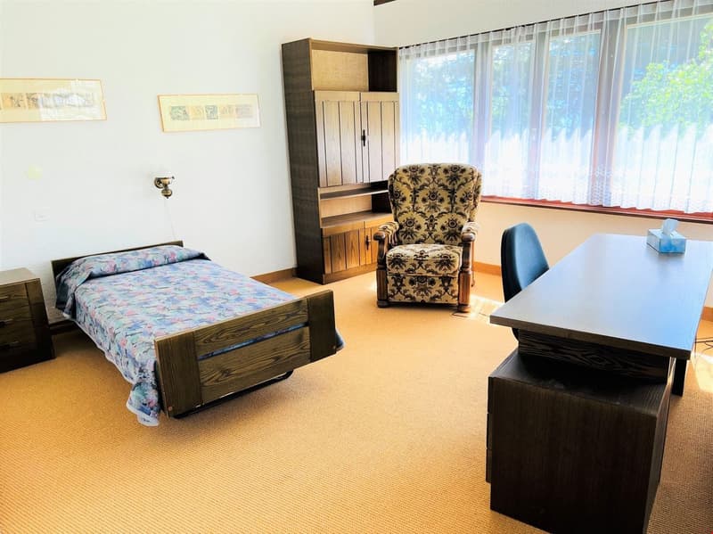 The image shows a bedroom with a bed, a floral patterned armchair, a wooden cabinet, and a desk. The room has large windows that provide natural light and a cozy, comfortable atmosphere.