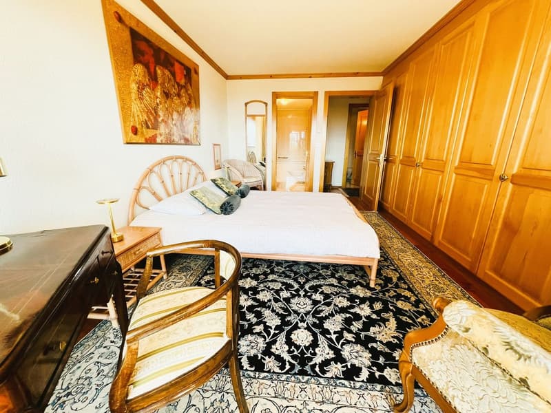 Bedroom with a bed, wooden bed frame, white bedding, decorative pillows, bedside tables, lamps, dresser, chair, and a wooden wardrobe.