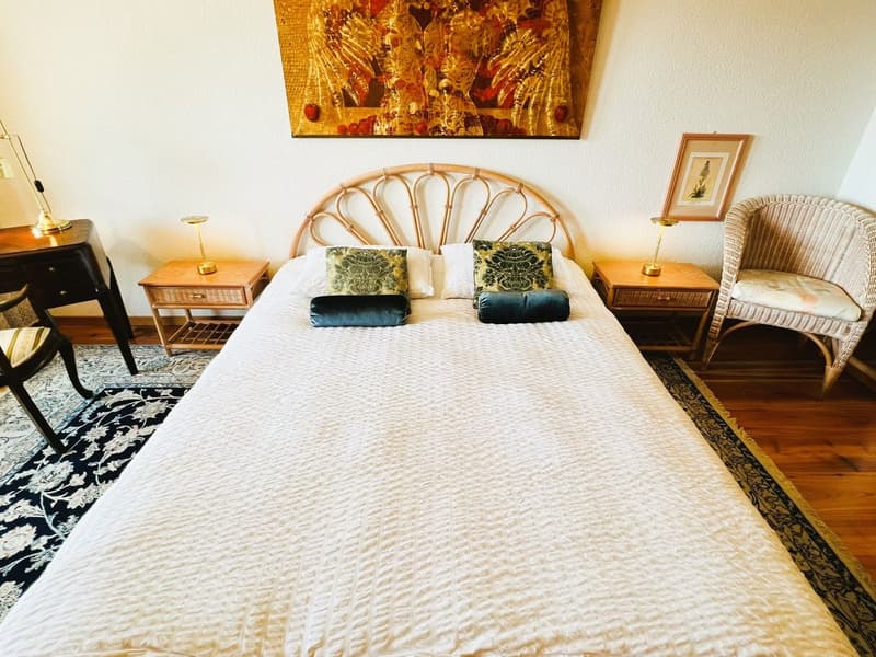 Rattan headboard, white bedspread, colorful pillows, night lamps, bedside tables, framed picture on the wall, wicker chair, dark rug, wooden floor, large tapestry