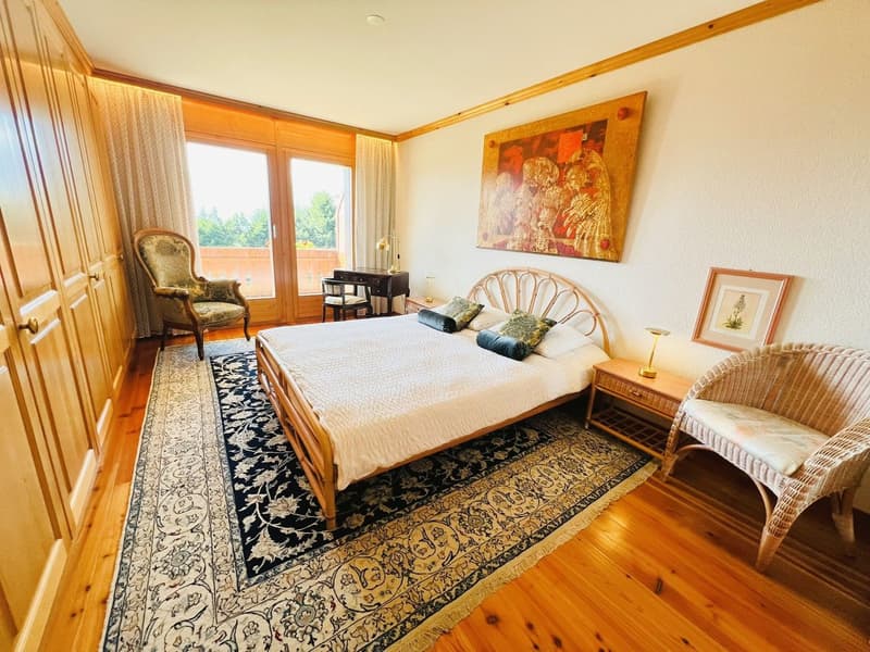Large bedroom with double bed, wooden bedframe, multiple pillows, bedside tables with lamps, wooden desk with chair, large window with balcony view, large rug, wooden armoire, artwork on wall.