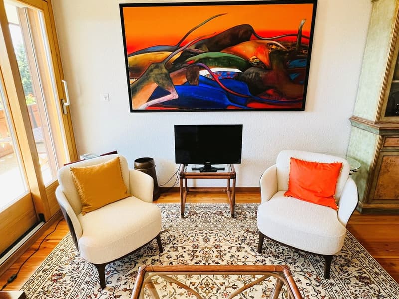 Two armchairs, a carpet, a coffee table, a flatscreen TV, a side table, an abstract painting on the wall, a wooden door, a balcony outside
