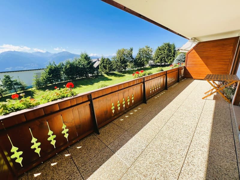 Wooden balcony, tile floor, wooden fence, table with folding legs, greenery and flowers on the railing, green lawn, trees, house, mountains in the distance, blue sky